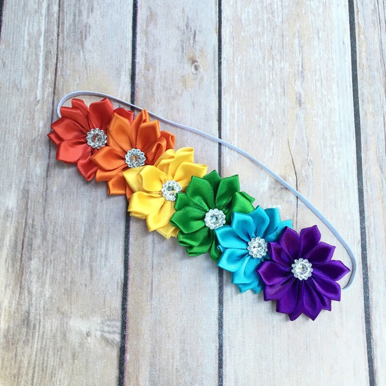 Puede incluir: Diadema arco&iacute;ris con seis flores de sat&eacute;n, cada una con un centro de pedrer&iacute;a. Las flores son rojas, naranjas, amarillas, verdes, azules y moradas.