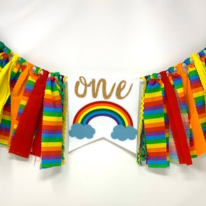 May include: A colorful banner with fabric strips in rainbow colors. A white pennant in the center features a rainbow, blue clouds, and the word "one" in gold glitter. Perfect for a first birthday celebration.