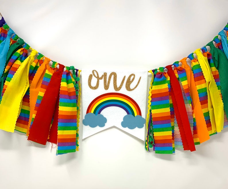 May include: A colorful banner with fabric strips in rainbow colors. A white pennant in the center features a rainbow, blue clouds, and the word "one" in gold glitter. Perfect for a first birthday celebration.