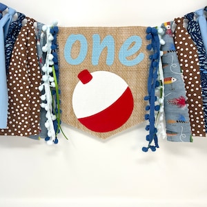 May include: A burlap banner with the word "one" in blue letters and a red and white fishing bobber. The banner is decorated with blue and white pom-poms, brown and white polka dot fabric, and blue and white striped fabric.