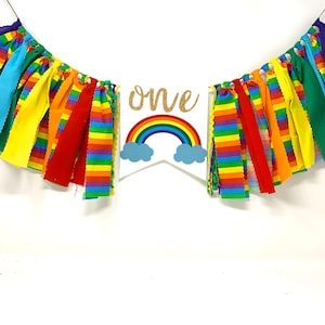 May include: A colorful rainbow-themed banner with the word "one" in gold glitter. The banner features a rainbow and cloud design, with strips of fabric in various colors, perfect for a birthday celebration.
