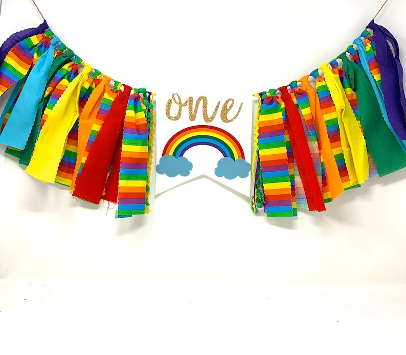 May include: A colorful rainbow-themed banner with the word "one" in gold glitter. The banner features a rainbow and cloud design, with strips of fabric in various colors, perfect for a birthday celebration.