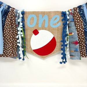 May include: A burlap banner with the word "one" in blue letters and a red and white fishing bobber. The banner is decorated with blue and white pom-poms, brown and white polka dot fabric, and blue and white striped fabric.