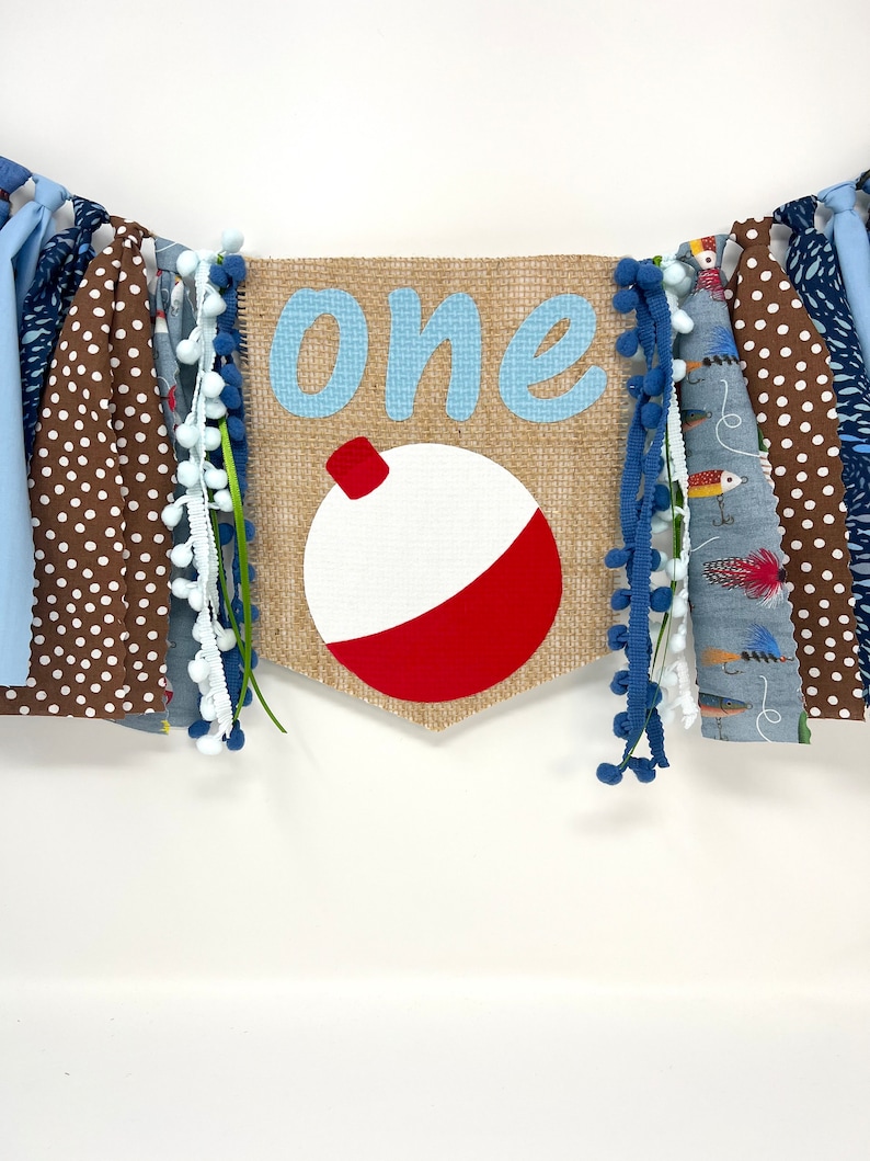 May include: A burlap banner with the word "one" in blue letters and a red and white fishing bobber. The banner is decorated with blue and white pom-poms, brown and white polka dot fabric, and blue and white striped fabric.
