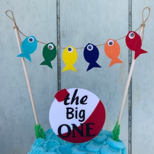 May include: A blue and white cake with a fishing-themed cake topper. The topper features a red and white circle with the text "the Big ONE" and a string of colorful felt fish with googly eyes.