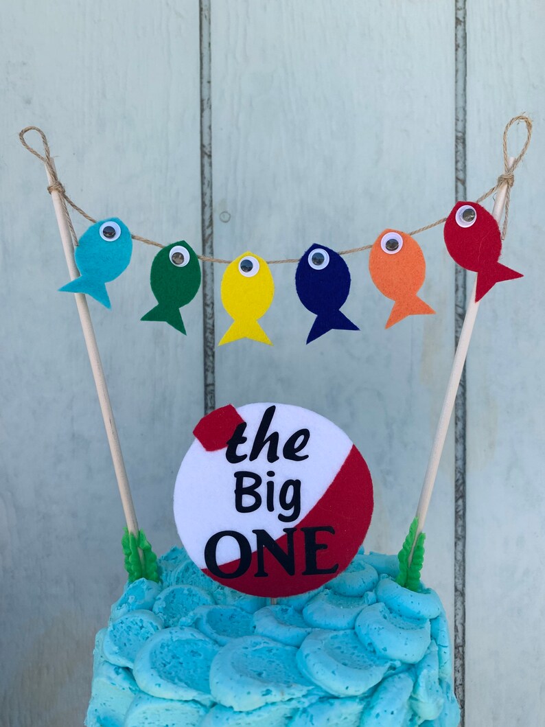May include: A blue and white cake with a fishing-themed cake topper. The topper features a red and white circle with the text "the Big ONE" and a string of colorful felt fish with googly eyes.