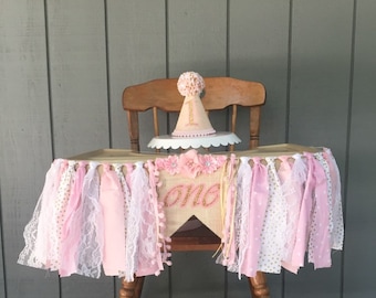 May include: A pink and gold high chair banner with the word "one" in gold glitter. The banner is made of burlap, lace, and ribbon. There is a pink and gold party hat on top of a cake on the high chair.