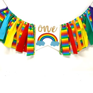 May include: A colorful rainbow-themed banner with the word "one" in gold glitter. The banner features a rainbow and cloud design, with strips of fabric in various colors. Perfect for a first birthday celebration.
