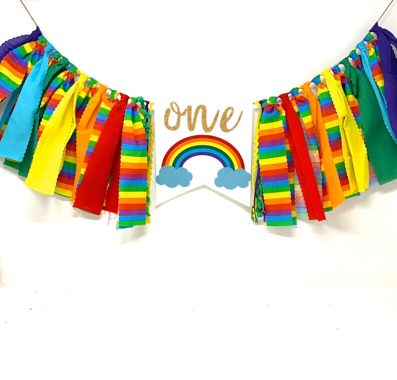 May include: A colorful rainbow-themed banner with the word "one" in gold glitter. The banner features a rainbow and cloud design, with strips of fabric in various colors. Perfect for a first birthday celebration.
