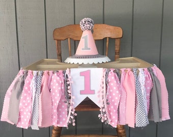 May include: A pink and gray high chair banner with a large white number one and pom poms. A matching pink and gray party hat with a white number one is on top of the high chair.