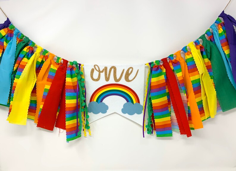 May include: A colorful rainbow-themed banner with the word "ONE" in gold glitter. The banner features a rainbow and cloud design, with a variety of colorful fabric strips in rainbow colors. The banner is perfect for a first birthday celebration.