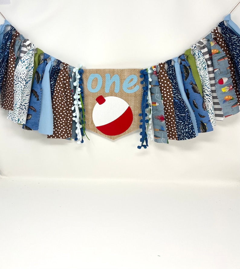 May include: A blue and brown fabric banner with the word "one" and a red and white fishing bobber on a burlap background. The banner is decorated with blue and white pom-poms.