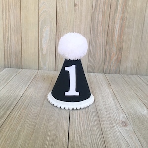 May include: A black and white party hat with the number 1 in white. The hat has a white pom-pom on top and a white scalloped trim at the base. The hat is sitting on a wooden surface.