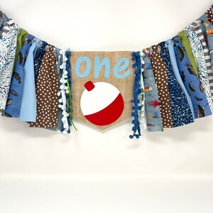 May include: A burlap banner with the word "one" in blue script and a red and white fishing bobber. The banner is decorated with blue, brown, and green fabric scraps.
