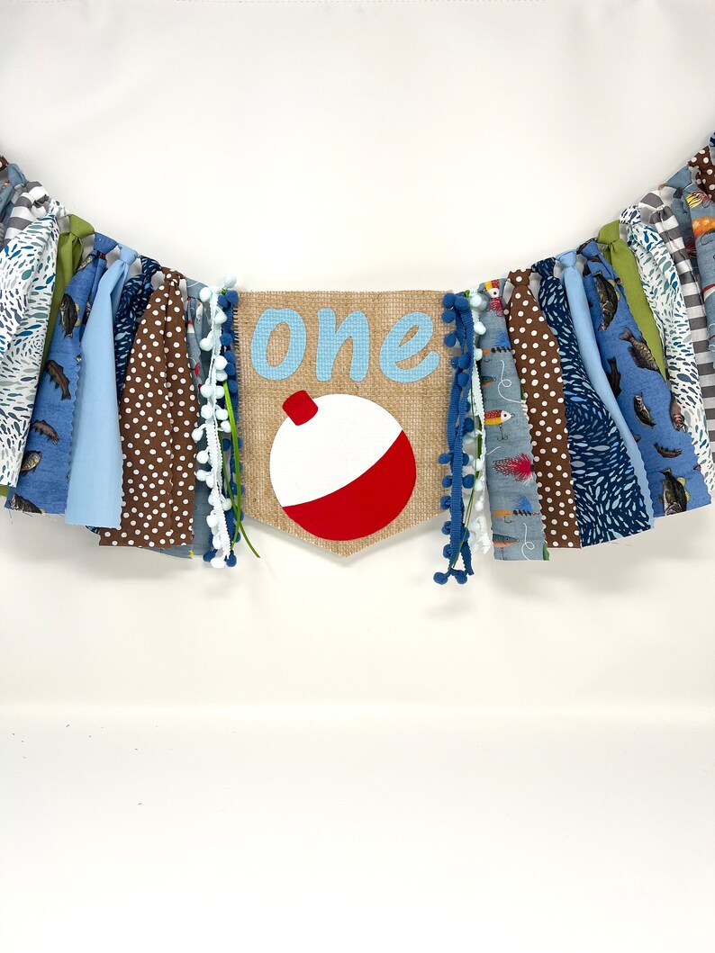 May include: A burlap banner with the word "one" in blue script and a red and white fishing bobber. The banner is decorated with blue, brown, and green fabric scraps.
