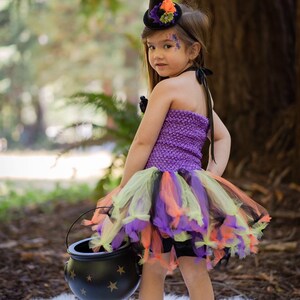 K&ouml;nnte beinhalten: Ein Kind in einem Halloween-Hexenkost&uuml;m. Das Outfit umfasst ein lila Strickoberteil, einen mehrlagigen T&uuml;llrock in Orange, Gr&uuml;n und Lila sowie einen kleinen schwarzen Hexenhut mit floralen Akzenten. Das Kind h&auml;lt einen schwarzen Kessel mit goldenen Sternen.