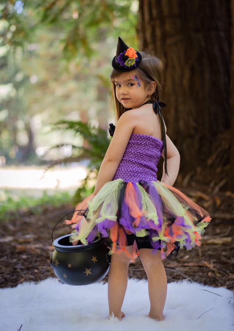 K&ouml;nnte beinhalten: Ein Kind in einem Halloween-Hexenkost&uuml;m. Das Outfit umfasst ein lila Strickoberteil, einen mehrlagigen T&uuml;llrock in Orange, Gr&uuml;n und Lila sowie einen kleinen schwarzen Hexenhut mit floralen Akzenten. Das Kind h&auml;lt einen schwarzen Kessel mit goldenen Sternen.