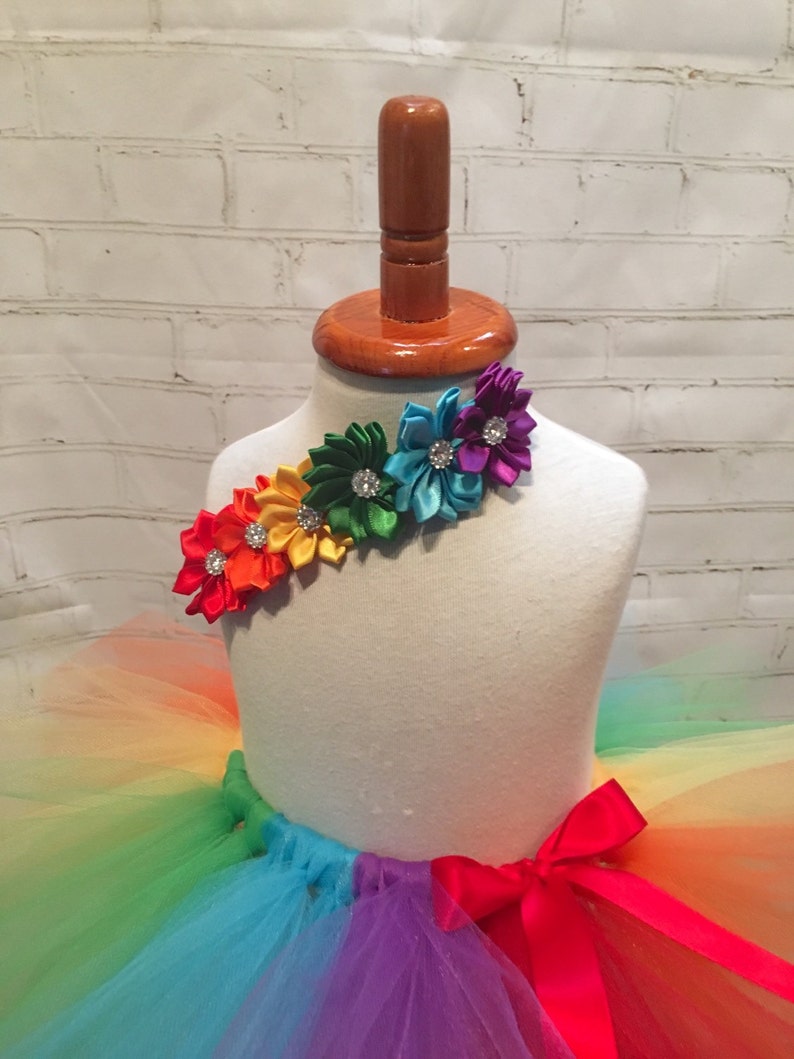 Puede incluir: Falda de tut&uacute; de tul arco&iacute;ris con un lazo de sat&eacute;n rojo y una corona de flores hecha de flores de cinta de sat&eacute;n de colores con pedrer&iacute;a.