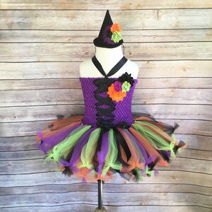 K&ouml;nnte beinhalten: Ein lila-schwarzes Halloween-Hexenkost&uuml;m mit passendem Hut. Das gestrickte lila Oberteil hat schwarze Banddetails. Der Tutu-Rock ist mit orangefarbenem, gr&uuml;nem, lila und schwarzem T&uuml;ll geschichtet. Der Hut ist schwarz mit orangefarbenen und gr&uuml;nen floralen Akzenten.