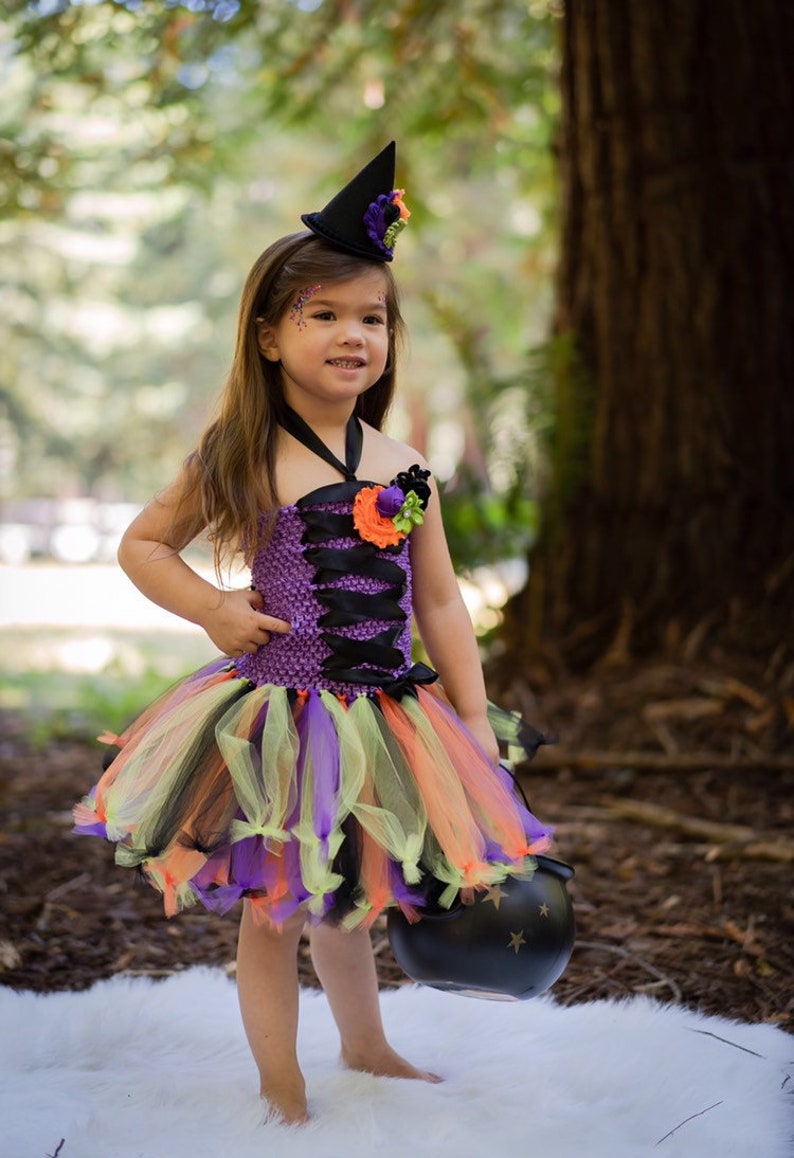 K&ouml;nnte beinhalten: Ein Kind in einem Halloween-Hexenkost&uuml;m. Das Outfit umfasst ein lila-schwarzes Oberteil, einen bunten Tutu-Rock und einen schwarzen Hexenhut. Das Kind h&auml;lt einen schwarzen Kessel mit Sternaussparungen.