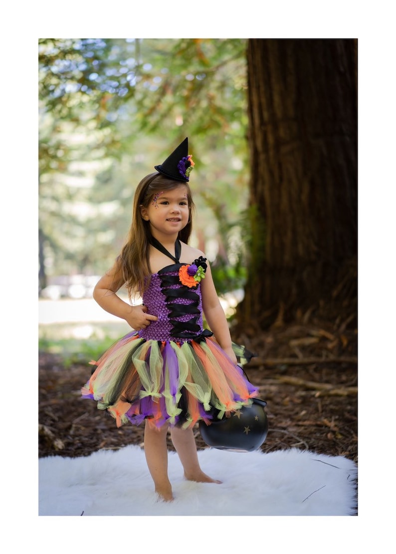 K&ouml;nnte beinhalten: Ein Halloween-Hexenkost&uuml;m f&uuml;r Kinder. Das Kleid ist lila, schwarz, orange und gr&uuml;n mit passendem Hut. Das Kind h&auml;lt einen schwarzen Kessel. Das Kost&uuml;m ist perfekt f&uuml;r das Trick-or-Treating.
