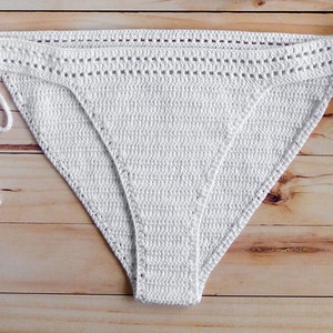 May include: White crocheted bikini bottom with adjustable ties on the sides. The bikini bottom is made of a delicate, open-weave fabric.
