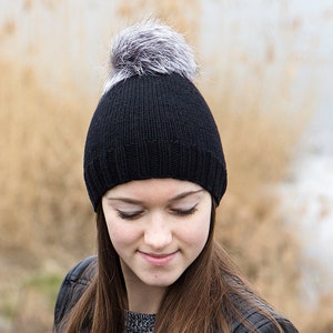 May include: A black knit beanie with a gray and white faux fur pom pom. The beanie is worn by a person with long brown hair.