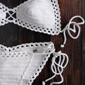 May include: A white crochet bikini set, featuring a triangle top and a low-rise bottom. The edges are detailed with a decorative openwork pattern. The bikini has adjustable ties and is displayed on a dark wood surface.
