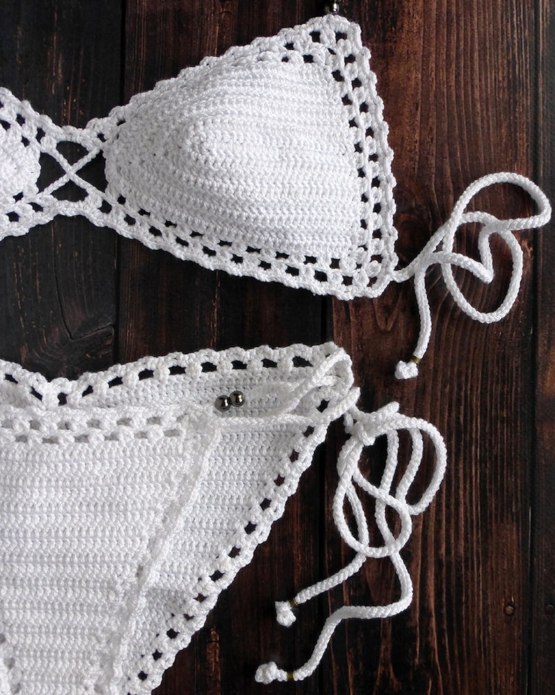 May include: A white crochet bikini set, featuring a triangle top and a low-rise bottom. The edges are detailed with a decorative openwork pattern. The bikini has adjustable ties and is displayed on a dark wood surface.