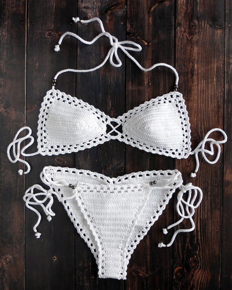 May include: A white, handmade crochet bikini set. The top features triangle cups with a criss-cross design, and the bottoms are a classic bikini cut. The set has adjustable ties and is displayed on a dark wood surface.
