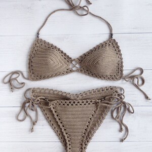 May include: A two-piece, taupe-colored crochet bikini set. The top features triangle cups with a criss-cross design and halter straps. The bottoms have a low-rise cut with adjustable side ties. The set is displayed on a white wooden surface.