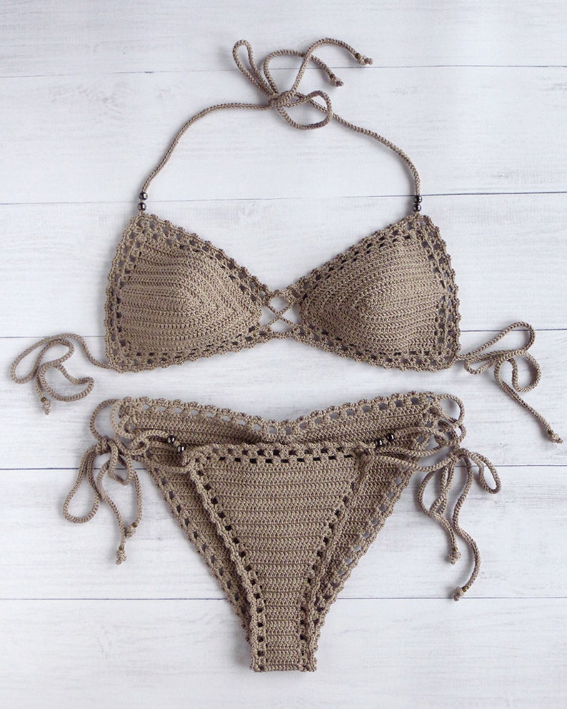 May include: A two-piece, taupe-colored crochet bikini set. The top features triangle cups with a criss-cross design and halter straps. The bottoms have a low-rise cut with adjustable side ties. The set is displayed on a white wooden surface.