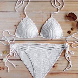 May include: A white two-piece crochet bikini set featuring a triangle top with scalloped edges and tie-up straps, paired with matching bikini bottoms that include side ties and a decorative open-stitch waistband detail for a textured finish.