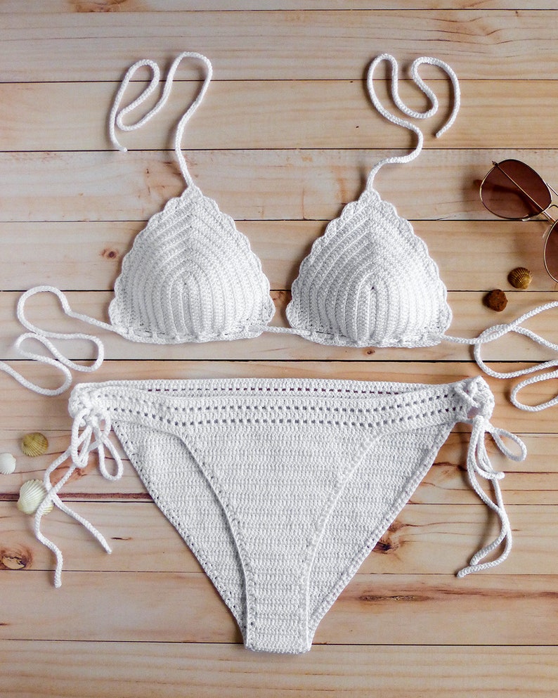 May include: A white two-piece crochet bikini set featuring a triangle top with scalloped edges and tie-up straps, paired with matching bikini bottoms that include side ties and a decorative open-stitch waistband detail for a textured finish.