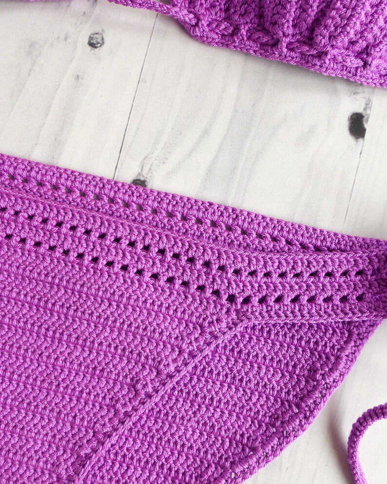 May include: A vibrant purple crochet bikini bottom featuring a dense, textured stitch pattern and a decorative horizontal band with openwork detailing along the upper edge, complemented by matching crochet ties at the side for a secure fit.