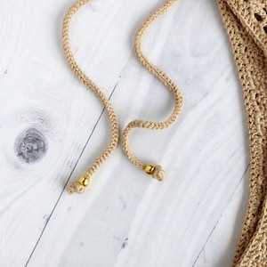 May include: Two beige cords with gold beads at the ends. The cords are on a white wooden surface.