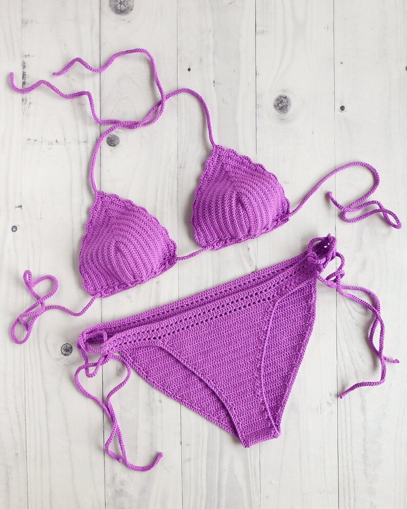 May include: A vibrant purple two-piece bikini set crafted from crochet yarn. The top features two triangular cups with scalloped edges and thin tie strings. The matching bottom piece includes side tie closures and a textured, ribbed crochet pattern.