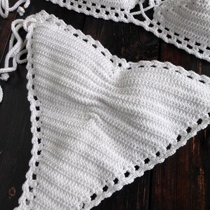 May include: A handmade white crochet bikini set. The top and bottom are displayed on a dark wood surface. The bikini top has triangle cups with adjustable ties. The edges of the bikini are detailed with a decorative openwork pattern.