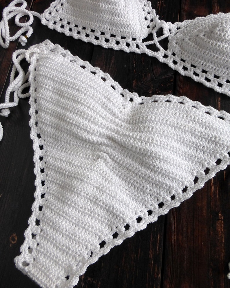 May include: A handmade white crochet bikini set. The top and bottom are displayed on a dark wood surface. The bikini top has triangle cups with adjustable ties. The edges of the bikini are detailed with a decorative openwork pattern.