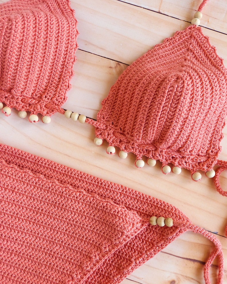 Puede incluir: Un conjunto de bikini de ganchillo color coral con textura acanalada en la parte superior triangular y la braga a juego. Los bordes de la parte superior del bikini est&aacute;n adornados con peque&ntilde;as cuentas de madera redondas y claras.