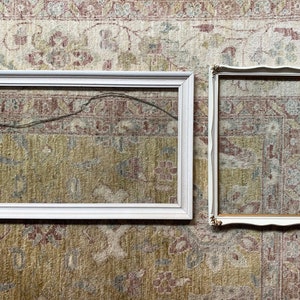 May include: Two empty picture frames. One is a large, rectangular frame with a simple white finish. The other is a smaller, ornate frame with a white finish and gold accents. Both frames are empty.