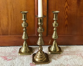 May include: Three gold metal candle holders with a square base and a tall, fluted design. A white candle is placed in the center holder.