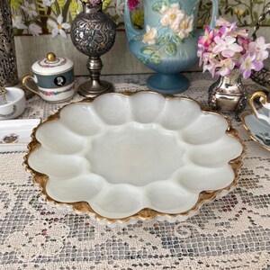 May include: A white, scalloped-edge serving dish with a gold rim. The dish has 12 individual compartments. The dish is on a lace tablecloth, with other decorative items in the background, including vases and flowers.
