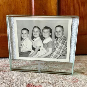 May include: A black and white photo of four children in a glass picture frame with a decorative border. The frame is sitting on a white rug with a pink floral pattern.