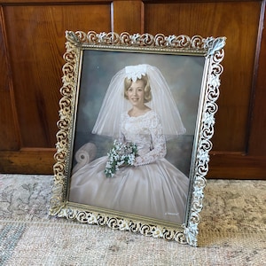 May include: A vintage gold-toned picture frame with an ornate design. The frame holds a black and white photograph of a bride wearing a white dress and veil. The bride is holding a bouquet of flowers.