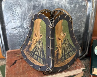 May include: A bell-shaped lampshade featuring black leather-like panels laced together with white cord. Each panel displays a printed illustration of two figures in long dresses standing near curtains in shades of green, yellow, and brown.