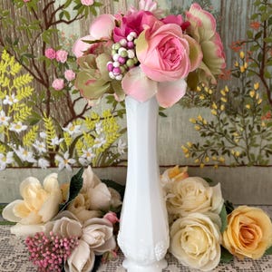 Peut inclure: Un vase blanc de style vintage rempli de fleurs artificielles roses et vertes. D'autres fleurs dans les tons de rose, cr&egrave;me et jaune sont dispos&eacute;es autour du vase. L'arri&egrave;re-plan pr&eacute;sente une tapisserie avec des motifs floraux et d'arbres.