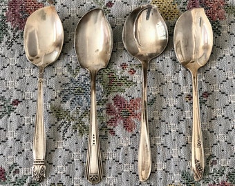 May include: Four silver spoons with ornate handles. The spoons are arranged in a row on a patterned fabric background.