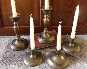 May include: Five brass candle holders with white candles. The candle holders are on a rug with a stack of books in the center.