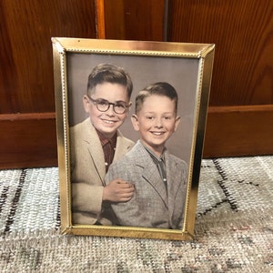 May include: A framed black and white photograph of two smiling boys in suits. The gold-colored frame surrounds the portrait, which is set against a neutral background. One boy wears glasses and a light-colored suit, while the other has a gray suit.